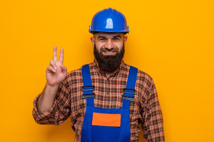Foto grátis construtor barbudo, com uniforme de construção e capacete de segurança, olhando para a câmera feliz e positivo, mostrando o sinal v sorrindo em pé sobre um fundo laranja