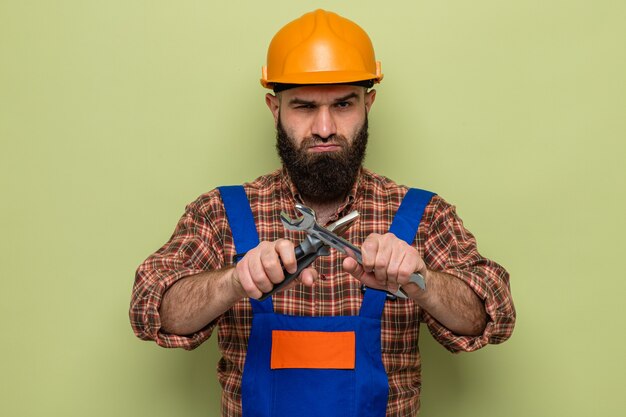 Construtor barbudo com uniforme de construção e capacete de segurança segurando a chave inglesa e um alicate, olhando para a câmera com o rosto sério, cruzando as mãos em pé sobre o fundo verde