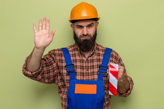 Construtor barbudo com uniforme de construção e capacete de segurança segurando fita adesiva, olhando com uma cara séria, fazendo gesto de parada com a mão