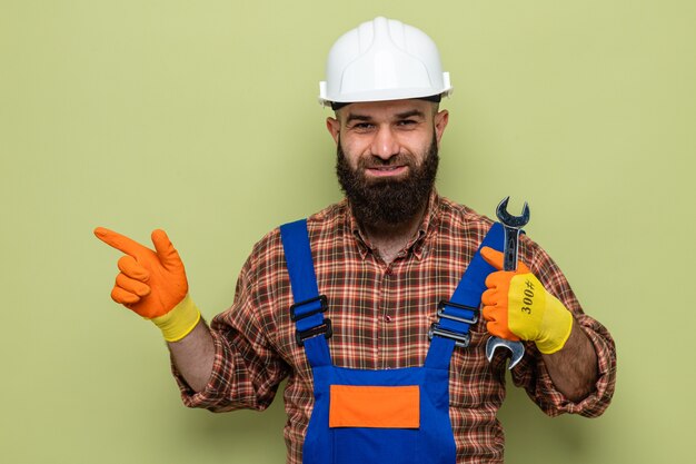 Construtor barbudo com uniforme de construção e capacete de segurança usando luvas de borracha segurando a chave inglesa olhando sorrindo apontando com o dedo indicador para o lado