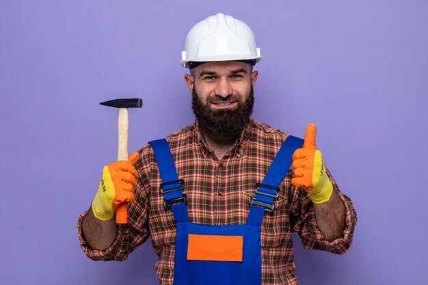 Construtor barbudo com uniforme de construção e capacete de segurança, usando luvas de borracha, segurando um martelo, parecendo sorridente e confiante mostrando os polegares para cima