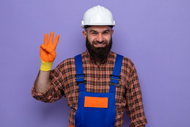 Construtor barbudo com uniforme de construção e capacete de segurança usando luvas de borracha, sorrindo alegremente mostrando o número três com os dedos