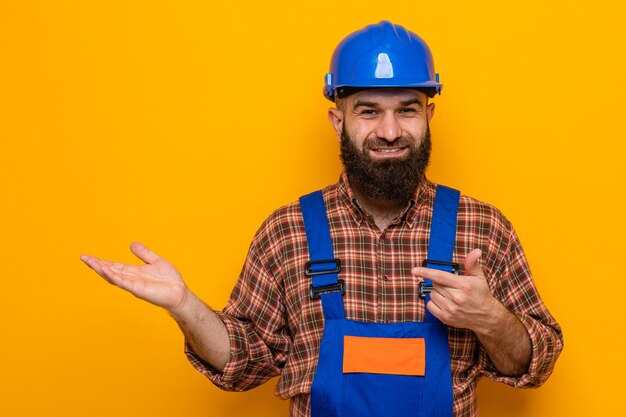 Construtor barbudo em uniforme de construção e capacete de segurança sorrindo alegremente apresentando algo com o braço de sua mão apontando com o dedo indicador para o lado