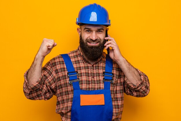 Construtor barbudo homem com uniforme de construção e capacete de segurança, punho cerrado feliz e animado, sorrindo enquanto fala ao telefone móvel em pé sobre um fundo laranja