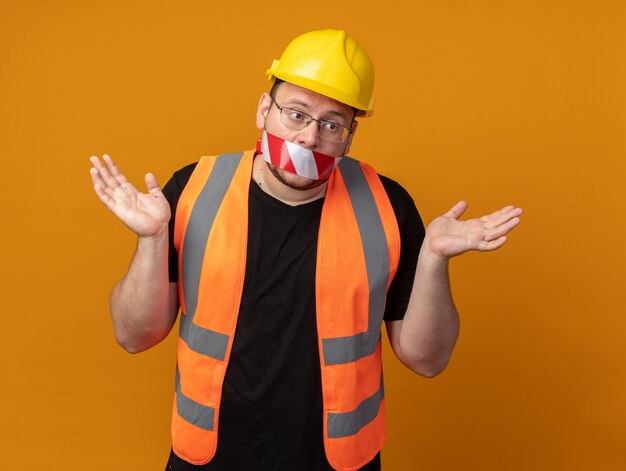 Construtor com colete de construção e capacete de segurança com fita adesiva sobre a boca, parecendo confuso, abrindo os braços para os lados