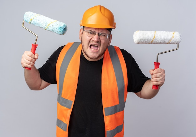 Foto grátis construtor furioso com colete de construção e capacete de segurança segurando rolos de tinta, gritando com expressão agressiva