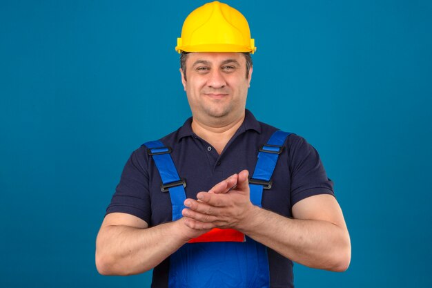 Construtor homem envelhecido médio vestindo uniforme de construção e capacete de segurança, esfregando as palmas das mãos e sorrindo olhando confiante sobre parede azul isolada