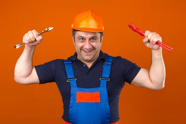 Foto grátis construtor homem vestindo uniforme de construção e capacete de segurança em pé com chaves nas mãos levantadas com sorriso no rosto sobre parede laranja isolada