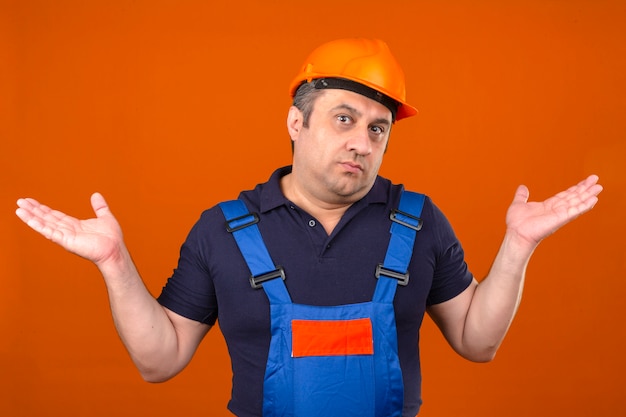 Construtor homem vestindo uniforme de construção e capacete de segurança, encolher os ombros os ombros espalhando as mãos à nora e expressão confusa sobre parede laranja isolada