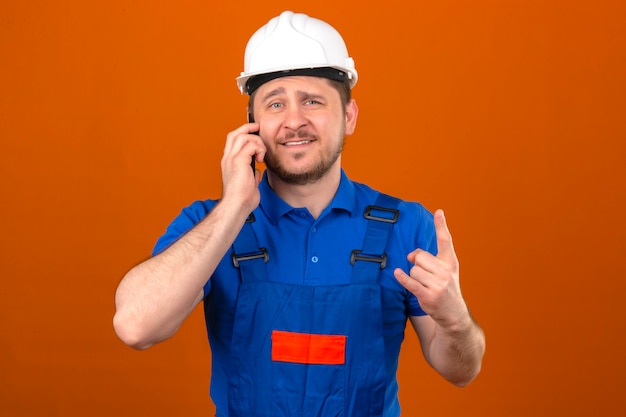 Foto grátis construtor homem vestindo uniforme de construção e capacete de segurança falando no celular mostrando o símbolo de rocha sorrindo alegremente em pé sobre a parede laranja isolada