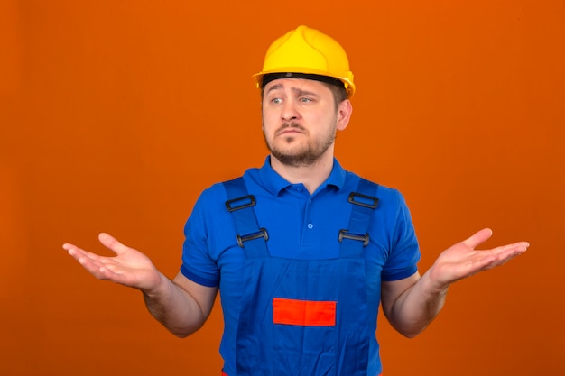 Construtor homem vestindo uniforme de construção e capacete de segurança sem noção e expressão confusa com braços e mãos levantadas conceito de dúvida sobre parede laranja isolada