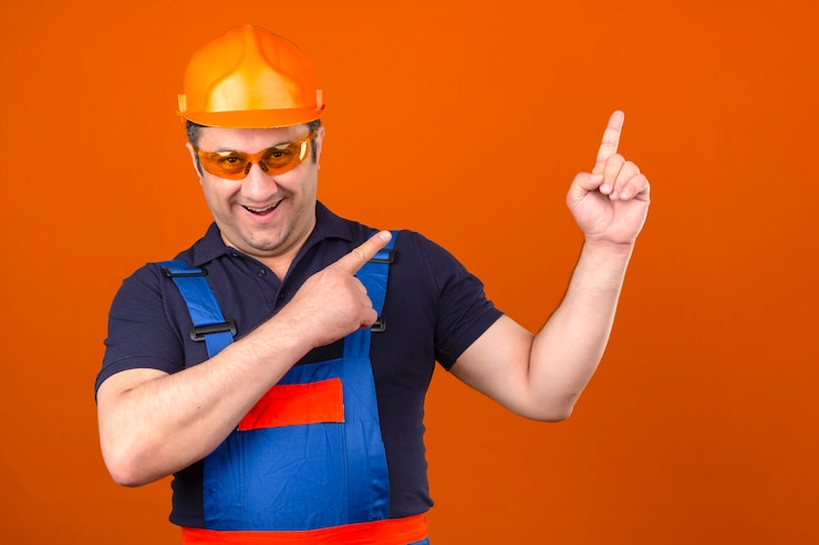 Foto grátis construtor homem vestindo uniforme de construção e capacete de segurança, sorrindo com cara feliz, apontando com as duas mãos e dedos para o lado ao longo da parede laranja isolada