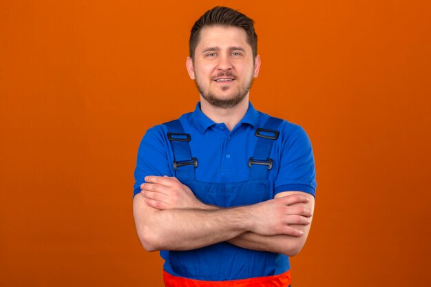 Construtor homem vestindo uniforme de construção em pé com os braços cruzados com sorriso confiante sobre parede laranja isolada