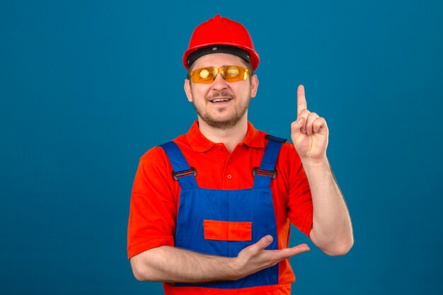 Construtor homem vestindo uniforme de construção óculos e capacete de segurança, apontando com o dedo, olhando confiante sorrindo novo conceito de idéia sobre parede azul isolada