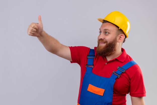 Construtor jovem barbudo com uniforme de construção e capacete de segurança olhando para o lado sorrindo mostrando os polegares para cima