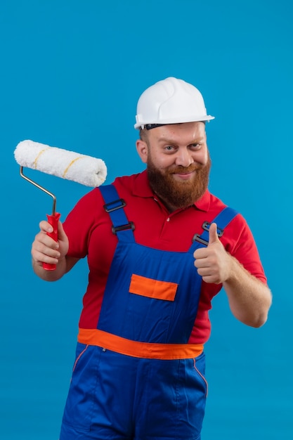 Construtor jovem barbudo com uniforme de construção e capacete de segurança segurando o rolo de pintura, sorrindo confiante mostrando os polegares para cima