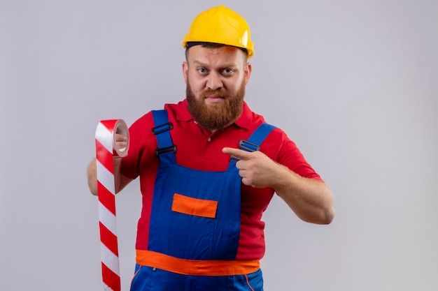 Construtor jovem barbudo, com uniforme de construção e capacete de segurança, segurando uma fita adesiva apontando com o dedo para ela, parecendo descontente