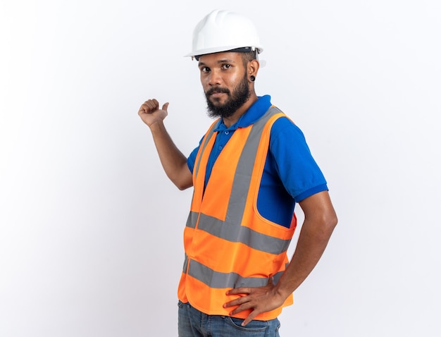 Foto grátis construtor jovem confiante de uniforme com capacete de segurança apontando para trás, isolado na parede branca com espaço de cópia