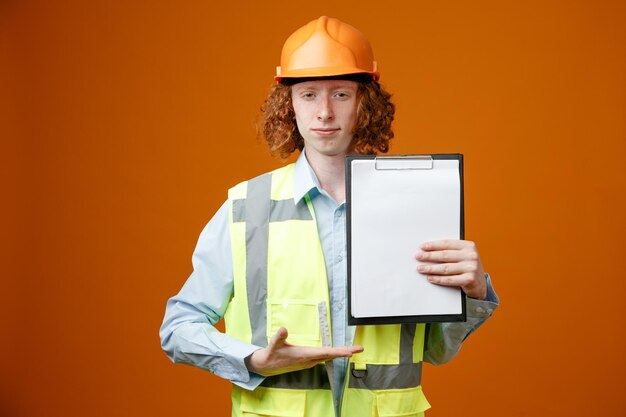 Construtor jovem em uniforme de construção e capacete de segurança apresentando prancheta com páginas em branco parecendo confiante em pé sobre fundo laranja