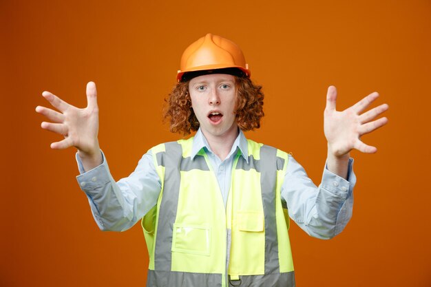 Construtor jovem em uniforme de construção e capacete de segurança olhando para a câmera sendo confuso e surpreso levantando os braços em pé sobre fundo laranja