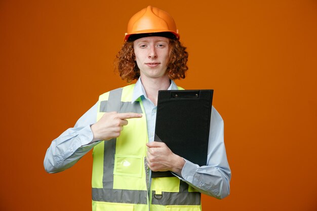 Construtor jovem em uniforme de construção e capacete de segurança segurando a área de transferência apontando com o dedo indicador para ele parecendo confiante em pé sobre fundo laranja