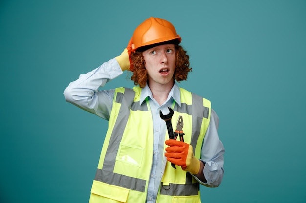 Construtor jovem em uniforme de construção e capacete de segurança segurando chave e alicate parecendo confuso com a mão na cabeça por erro em pé sobre fundo azul
