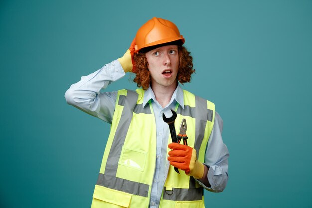 Construtor jovem em uniforme de construção e capacete de segurança segurando chave e alicate parecendo confuso com a mão na cabeça por erro em pé sobre fundo azul