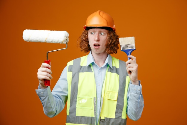 Construtor jovem em uniforme de construção e capacete de segurança segurando o rolo de pintura e pincel parecendo confuso com dúvidas sobre fundo laranja