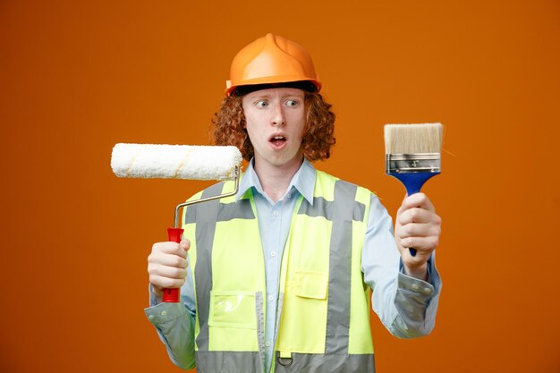 Construtor jovem em uniforme de construção e capacete de segurança segurando o rolo de pintura e pincel parecendo confuso com dúvidas sobre fundo laranja