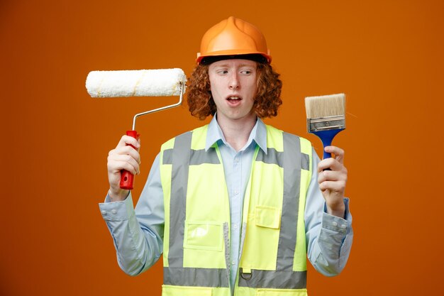 Construtor jovem em uniforme de construção e capacete de segurança segurando o rolo de pintura e pincel parecendo confuso com dúvidas sobre fundo laranja