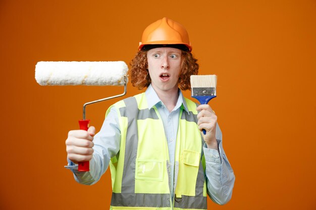 Construtor jovem em uniforme de construção e capacete de segurança segurando o rolo de pintura e pincel parecendo confuso com dúvidas sobre fundo laranja