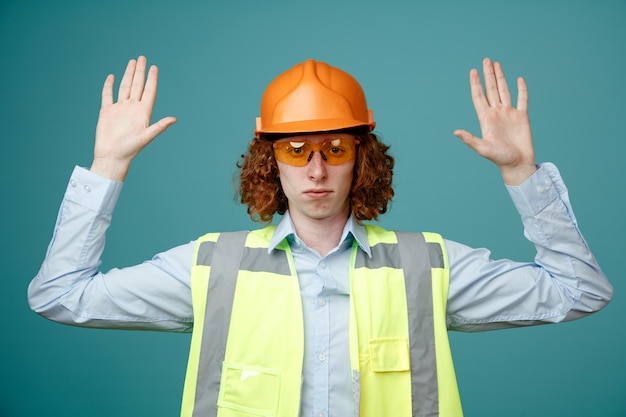 Construtor jovem em uniforme de construção e capacete de segurança usando óculos de segurança, olhando para a câmera com cara séria, levantando os braços sobre fundo azul