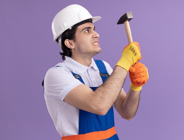 Construtor jovem furioso com uniforme de construção e capacete de segurança com luvas de borracha segurando um martelo olhando para ele com expressão irritada em pé sobre a parede roxa
