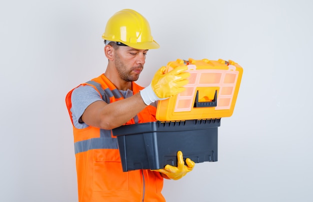 Construtor masculino abrindo caixa de ferramentas de plástico de uniforme, capacete, luvas, vista frontal.