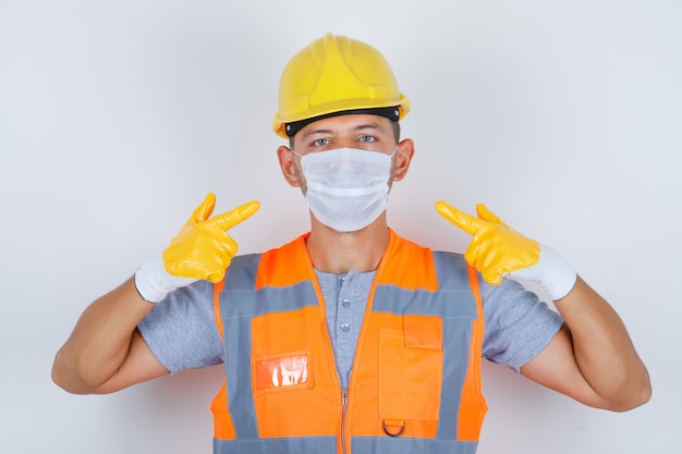 Construtor masculino apontando sua máscara médica de uniforme, capacete, luvas, vista frontal