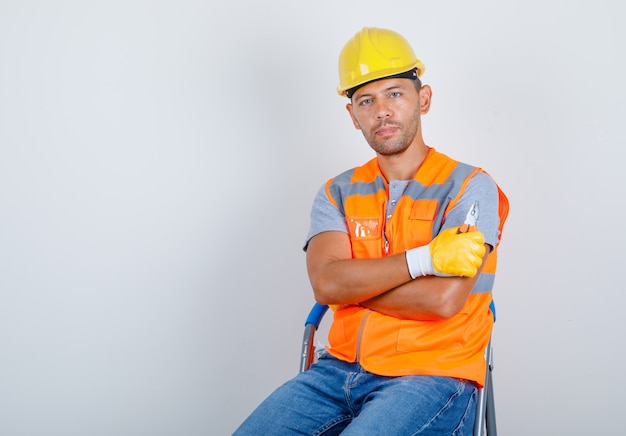 Construtor masculino de uniforme, jeans, capacete, luvas, sentado com os braços cruzados, vista frontal.