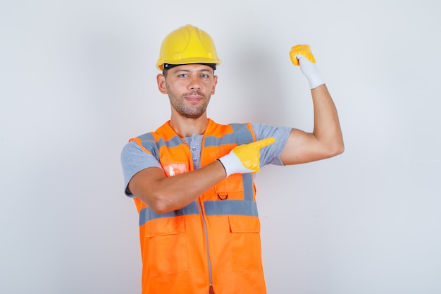 Foto grátis construtor masculino de uniforme, mostrando seu bíceps com o dedo e parecendo confiante, vista frontal.