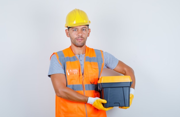 Construtor masculino segurando a caixa de ferramentas de uniforme, capacete, luvas, vista frontal.