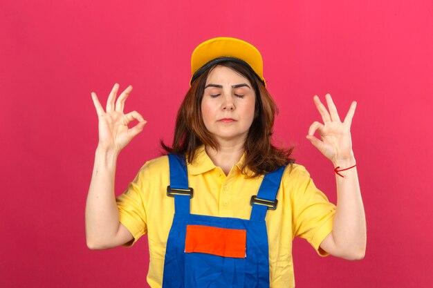 Construtor mulher vestindo uniforme de construção e boné amarelo relaxar e sorrindo com os olhos fechados, fazendo o gesto de meditação com os dedos sobre a parede rosa isolada