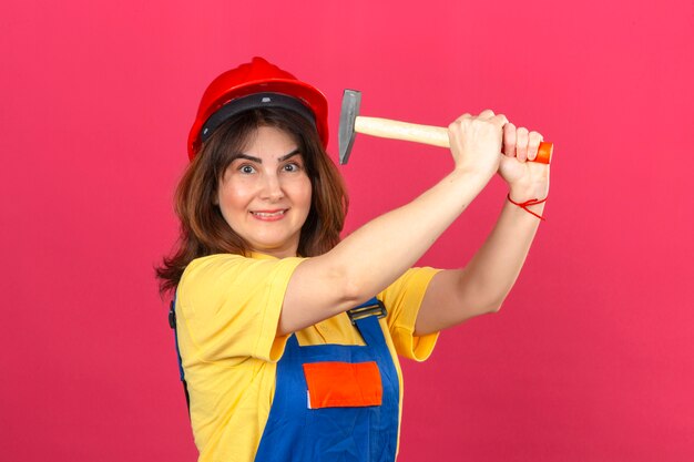 Construtor mulher vestindo uniforme de construção e capacete de segurança com sorriso ameaçando bater com martelo se divertindo sobre parede rosa isolada