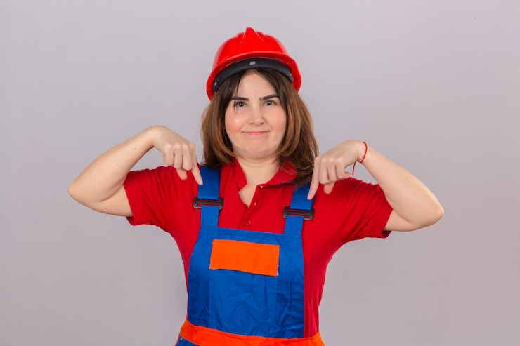Foto grátis construtor mulher vestindo uniforme de construção e capacete de segurança, olhando confiante com sorriso no rosto, apontando com os dedos para baixo sobre parede branca isolada