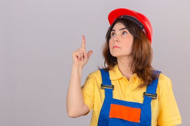 Foto grátis construtor mulher vestindo uniforme de construção e capacete de segurança, olhando para cima e batendo com o dedo para algo com interesse sobre parede branca isolada