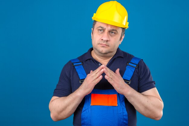 Construtor pensativo envelhecido médio homem vestindo uniforme de construção e capacete de segurança juntos de mãos dadas e pensando sobre isolado parede azul