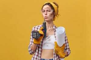 Construtora autoconfiante usando óculos de proteção, blusa branca e camisa quadriculada, luvas de proteção segurando a broca e os papéis sujos após um trabalho árduo isolado sobre a parede amarela. manutenção