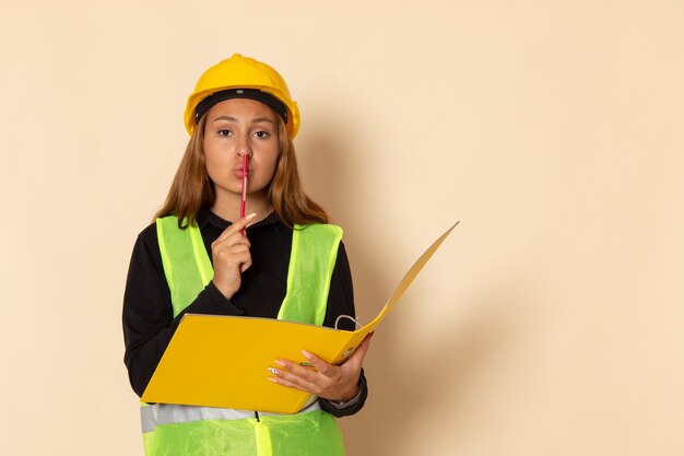 Construtora de frente com capacete amarelo segurando um documento amarelo e lápis na parede branca