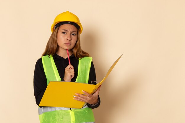 Construtora de frente com capacete amarelo segurando uma pasta amarela e pensando na parede branca