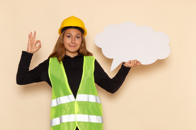 Construtora feminina de vista frontal com capacete amarelo segurando uma placa branca com um sorriso feminino na parede branca