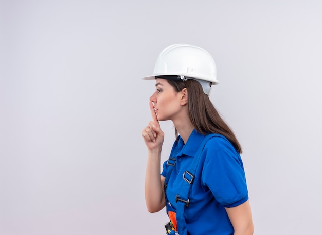 Construtora jovem confiante com capacete de segurança branco e uniforme azul fica de lado e faz gestos de silêncio no fundo branco isolado com espaço de cópia