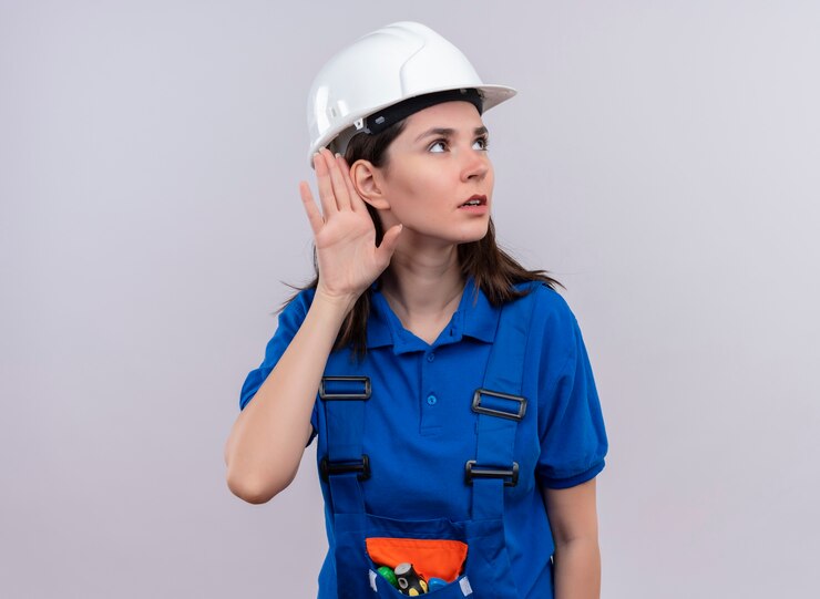 Foto grátis construtora jovem confiante com capacete de segurança branco e uniforme azul finge ouvir em um fundo branco isolado com espaço de cópia