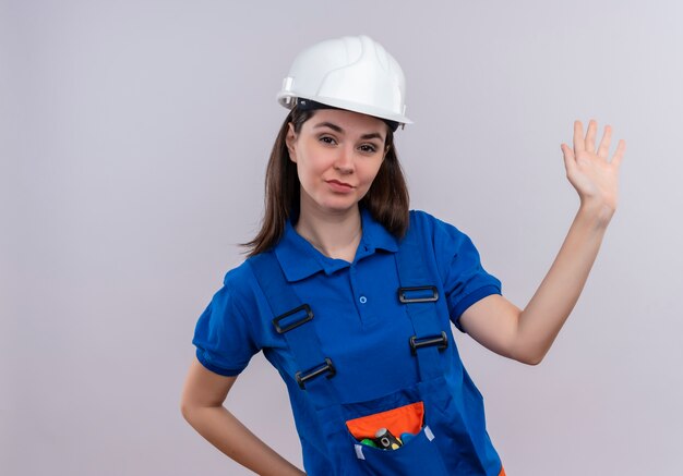 Construtora jovem confiante com capacete de segurança branco e uniforme azul levanta a mão e coloca a mão na cintura em um fundo branco isolado com espaço de cópia